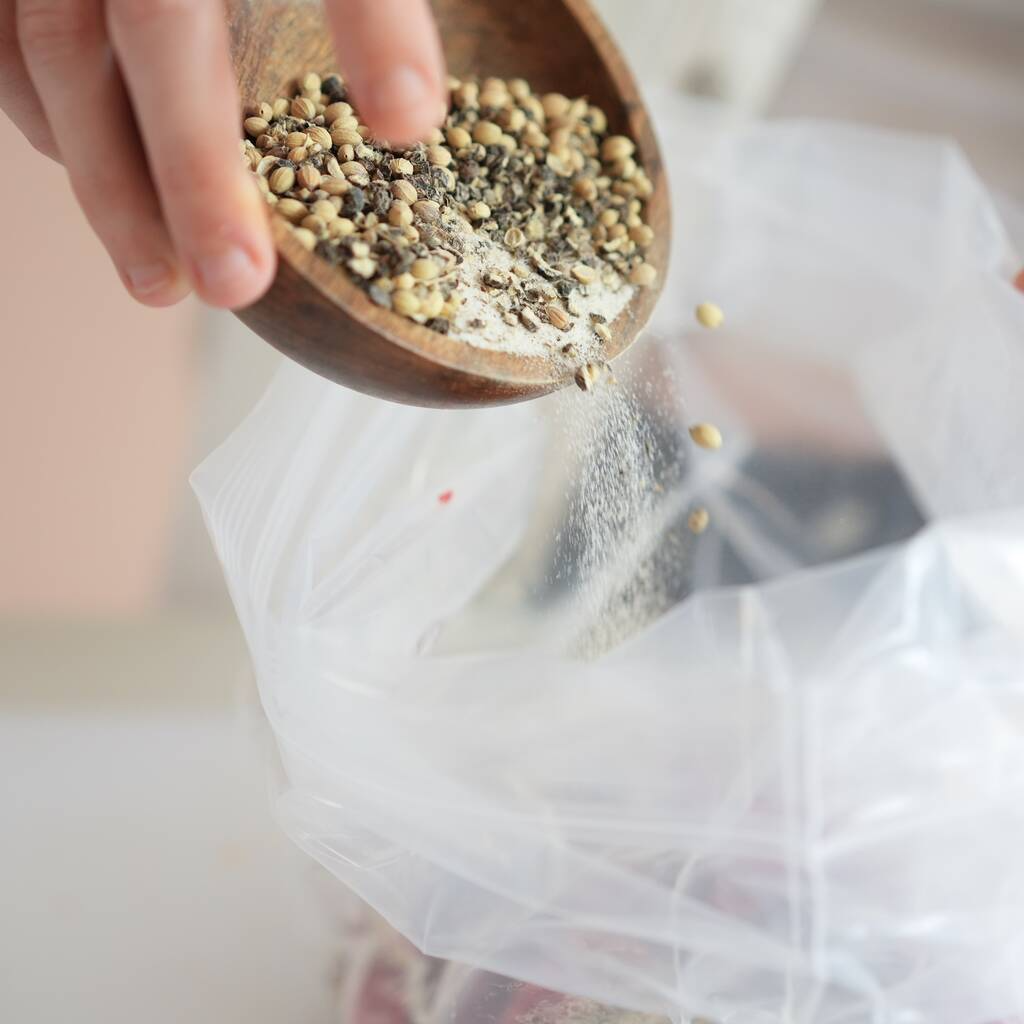 A person pouring various Sweet Chilli - Sliced Biltong spices from a wooden bowl into a clear plastic bag for seasoning.
